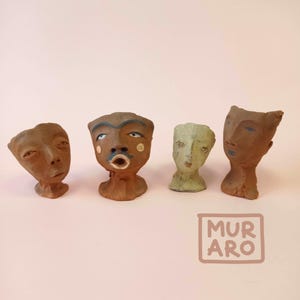 Puede incluir: Cuatro pequeñas esculturas de cerámica de rostros, cada una con características únicas y detalles pintados. Las esculturas son marrones, verdes y beige. La palabra "MURARO" está escrita en negro sobre un rectángulo blanco en la esquina inferior derecha de la imagen.