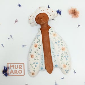 May include: A ceramic wall sculpture of a person wearing a white hat and a floral patterned dress. The sculpture is painted in shades of brown, white, blue, and peach.