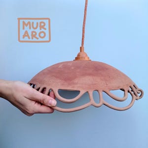 May include: A terracotta-colored ceramic pendant light with a decorative, scalloped edge. The light is suspended from a brown cord and has a brass socket.