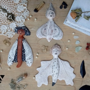 May include: Three handcrafted ceramic figures with stylised faces and wings, each featuring unique patterns and designs. The figures are in shades of white, brown, and black, arranged on a wooden surface with decorative elements like a feather and dried flowers.