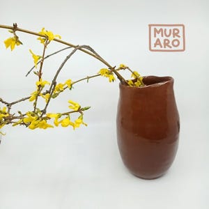 Peut inclure: Un vase en céramique brun avec une forme arrondie et un bord légèrement irrégulier. Le vase est vide et a une finition brillante. Une branche avec des fleurs jaunes est en arrière-plan.
