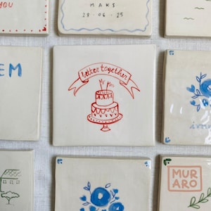 Könnte beinhalten: Quadratische Keramikfliesen mit handgezeichneten Designs. Eine Fliese zeigt einen roten Kuchen mit dem Schriftzug "better together" auf einem Banner. Andere Fliesen haben blaue Blumenmuster, Text und Daten. Die Fliesen sind auf einer weißen Oberfläche angeordnet.
