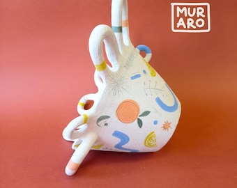 Puede incluir: Jarrón de cerámica blanca con un diseño abstracto único que presenta rodajas de naranja, hojas y formas geométricas en azul, amarillo y verde. El jarrón tiene tres asas y el texto "MURARO" es visible en el fondo.