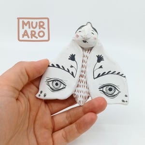 Könnte beinhalten: Weißer Keramik-Schmetterlings-Anhänger mit schwarzen Details, darunter Augen, Blumen und Ranken. Der Schmetterling hat einen rot-weiß gemusterten Körper.