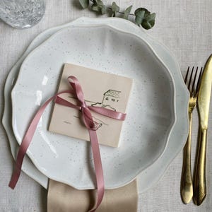 Könnte beinhalten: Ein weiß gesprenkeltes Keramik-Plattenset mit dekorativem Rand, eine kleine Karte mit rosa Band gebunden und goldenem Besteck. Die Karte zeigt eine einfache Hausillustration. Die Szene ist auf einer neutralen Leinentischdecke.