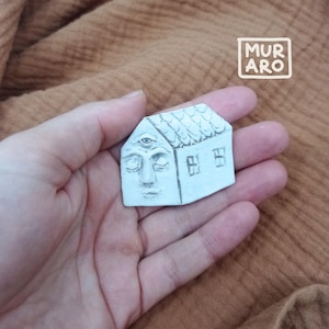 May include: White ceramic house shaped ornament with a closed eye face and a third eye on the forehead. The house has two windows and a roof with a textured design.