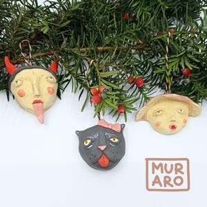 Op de afbeelding: Drie handgemaakte kerstversieringen van keramiek die aan een groene dennentak hangen. De versieringen zijn een zwarte kat met een roze strik, een geel gezicht met een roze hoed en een wit gezicht met rode hoorns. De versieringen hebben allemaal beschilderde details en steken hun tong uit.
