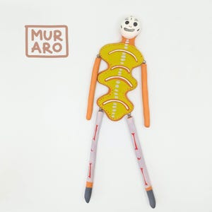 Puede incluir: Una muñeca esqueleto amarilla y naranja con una cabeza de calavera blanca. La muñeca tiene una sonrisa pintada y está articulada en los hombros, codos, caderas y rodillas. La muñeca está de pie sobre un fondo blanco. El texto "MURARO" está en un cuadrado en la esquina superior izquierda de la imagen.