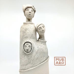 May include: White ceramic sculpture of three figures with abstract faces. The figures are stacked on top of each other, with the top figure wearing a hat. The sculpture is signed "MURARO" in the bottom right corner.