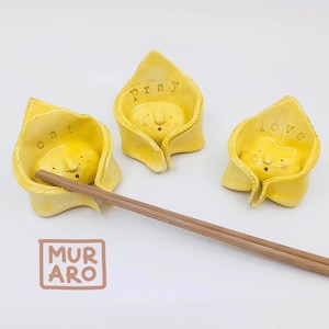 May include: Three yellow ceramic dumpling-shaped holders with smiling faces and the words "cat", "pray", and "love". A pair of wooden chopsticks rests in front of the holders. The logo "MURARO" is in a brown box.