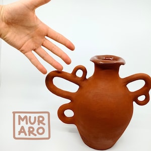May include: A terracotta vase with an abstract, organic shape and a glazed opening. The vase has multiple handles that resemble loops or swirls. The vase is sitting on a white surface. The text "MURARO" is written in black on a white background.
