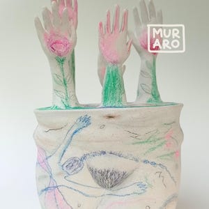 May include: A white ceramic vase with hand-drawn illustrations. The vase features multiple hand-shaped forms with pink and green accents, and the word "MURA RO" in a white square. The vase has a textured surface with additional drawings.