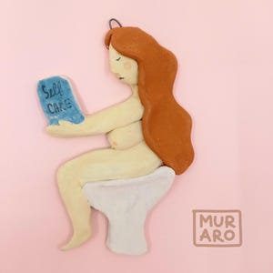 May include: A ceramic figurine of a person sitting on a toilet, reading a book that says "Self Care". The figurine has red hair and is wearing a white toilet seat. The background is pink.