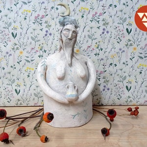 May include: White ceramic sculpture of a person with a crescent moon on their head, holding a small ceramic jug. The sculpture is standing on a wooden surface with a floral patterned background.