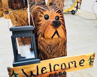 Puede incluir: Un oso de madera tallado sosteniendo una linterna y un letrero que dice "Welcome". El oso es marrón y tiene la nariz y los ojos negros. El letrero está hecho de madera y tiene un diseño de pino verde.