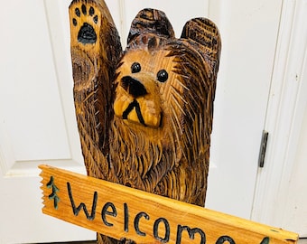Puede incluir: Una escultura de oso de madera tallada sosteniendo un cartel que dice "Bienvenido". El oso es marrón y tiene una huella de pata negra en su mano levantada. El cartel está hecho de madera y tiene un diseño de pino verde en cada extremo.