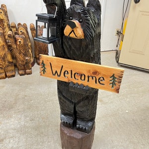 May include: A carved wooden black bear holding a lantern and a sign that says "Welcome". The bear is standing on a wooden base.