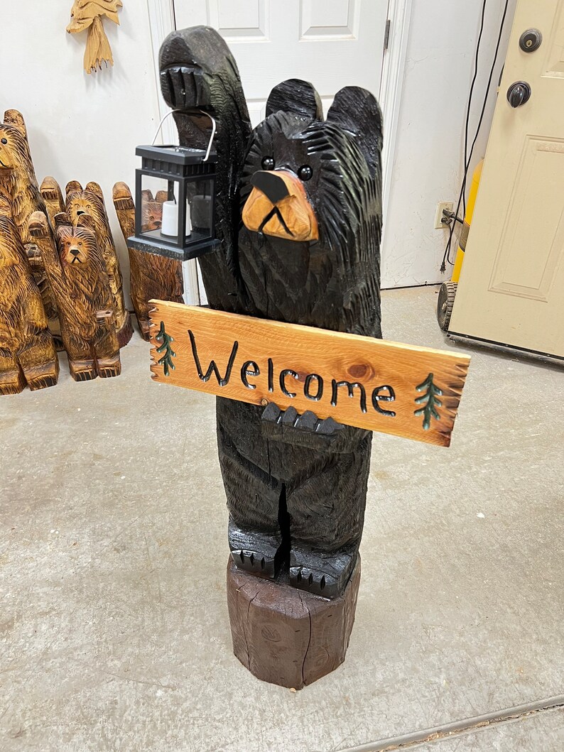 May include: A carved wooden black bear holding a lantern and a sign that says "Welcome". The bear is standing on a wooden base.
