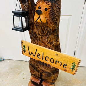 May include: A carved wooden bear sculpture holding a lantern and a sign that says "Welcome". The bear is brown and has a black nose and eyes. The sign is made of wood and has green pine trees painted on it.