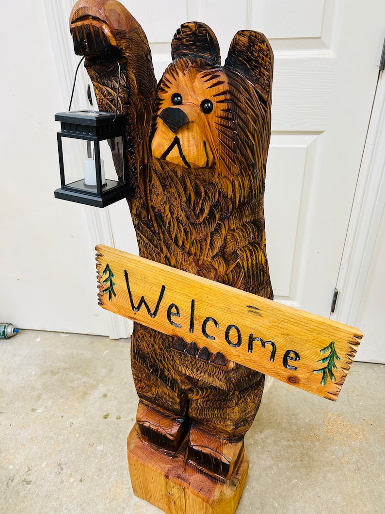 May include: A carved wooden bear sculpture holding a lantern and a sign that says "Welcome". The bear is brown and has a black nose and eyes. The sign is made of wood and has green pine trees painted on it.