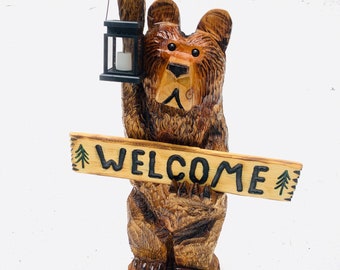 May include: A carved wooden bear holding a lantern and a sign that says "WELCOME". The bear is brown and has a black nose and eyes. The sign is made of wood and has a green pine tree design on each end.