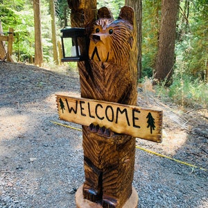 May include: A carved wooden bear sculpture holding a lantern and a sign that says "WELCOME". The bear is standing on a tree stump and is surrounded by trees.