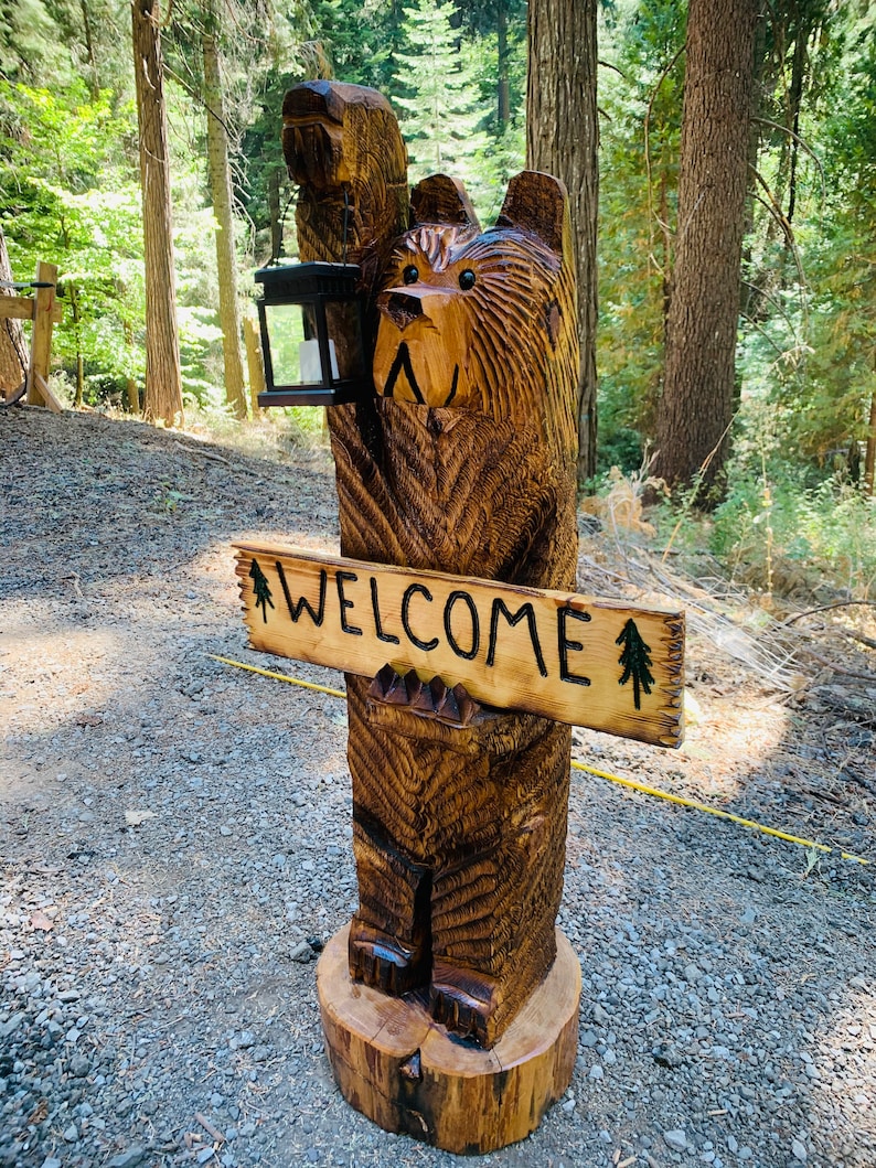 May include: A carved wooden bear sculpture holding a lantern and a sign that says "WELCOME". The bear is standing on a tree stump and is surrounded by trees.