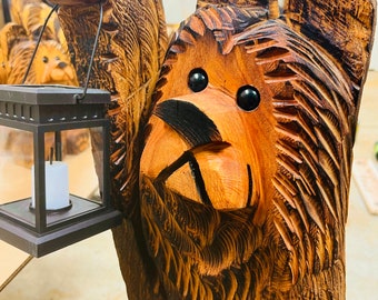 May include: A carved wooden bear sculpture with a lantern in its paw. The bear has a brown and black wood grain finish and realistic features, including eyes, a nose, and a mouth. The lantern is black with a glass pane and a white candle inside.
