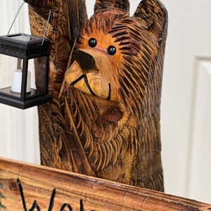 May include: A carved wooden bear sculpture holding a lantern with a welcome sign. The bear is brown with black eyes and a carved wood grain texture. The sign is made of wood and has the word "Welcome" written in black paint.