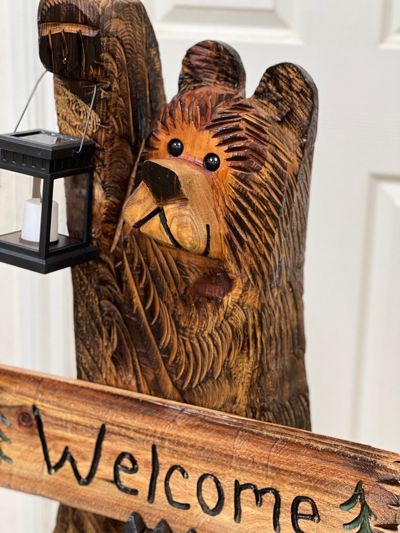 May include: A carved wooden bear sculpture holding a lantern with a welcome sign. The bear is brown with black eyes and a carved wood grain texture. The sign is made of wood and has the word "Welcome" written in black paint.