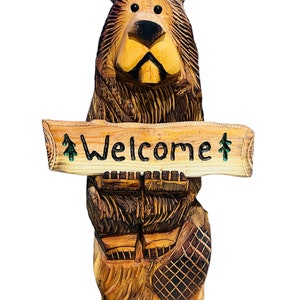 May include: A carved wooden bear holding a sign that says "Welcome". The bear is brown and has a black nose and eyes. The sign is made of wood and has a green pine tree on each end.