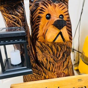 May include: A carved wooden bear sculpture with a solar-powered lantern and a sign that reads "Welcome". The bear is brown and has a black nose and eyes. The lantern is black and has a white candle inside. The sign is made of wood and has a green pine tree design.