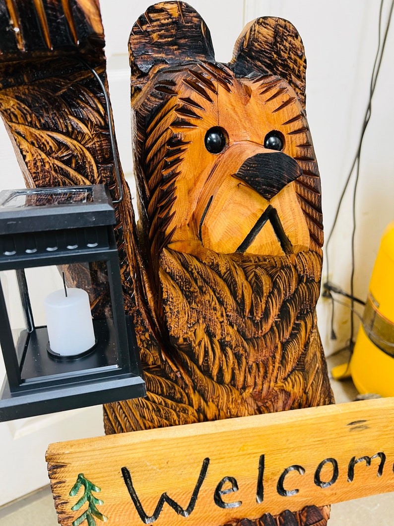 May include: A carved wooden bear sculpture with a solar-powered lantern and a sign that reads "Welcome". The bear is brown and has a black nose and eyes. The lantern is black and has a white candle inside. The sign is made of wood and has a green pine tree design.
