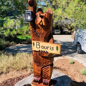 May include: A carved wooden bear holding a lantern and a sign that says "Bouris" with a small pine tree on each side of the text. The bear is standing on a tree stump.
