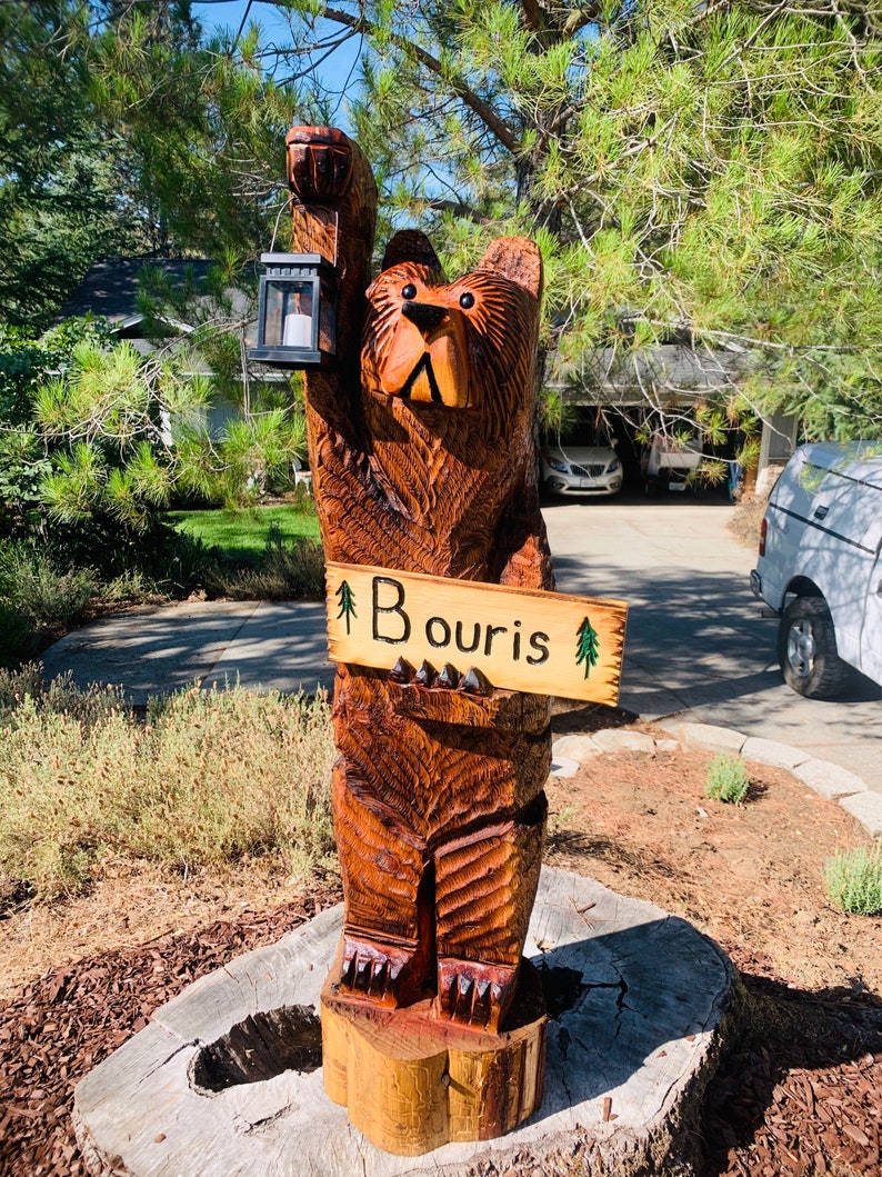 May include: A carved wooden bear holding a lantern and a sign that says "Bouris" with a small pine tree on each side of the text. The bear is standing on a tree stump.