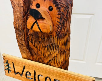 Puede incluir: Una escultura de oso de madera tallada sosteniendo un cartel que dice "Welcome" con pinos a cada lado del texto. El oso es marrón y tiene ojos negros y una nariz negra.