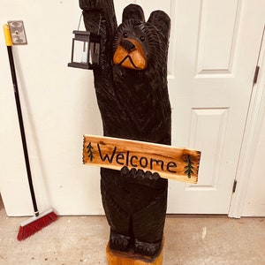 May include: A carved wooden black bear holding a lantern and a sign that says "Welcome". The bear is standing on a wooden base.