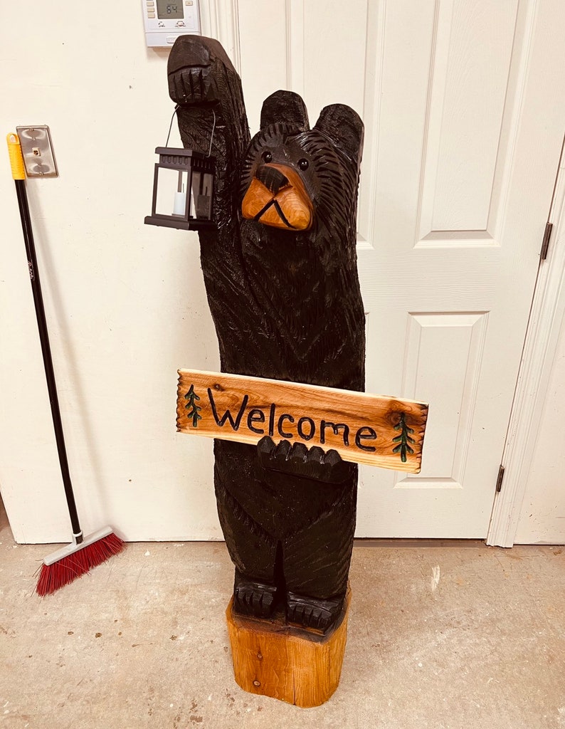 May include: A carved wooden black bear holding a lantern and a sign that says "Welcome". The bear is standing on a wooden base.