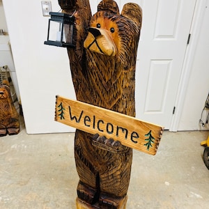 May include: A carved wooden bear statue holding a lantern and a sign that reads "Welcome." The bear is brown with detailed wood grain, standing on a wooden base. The sign has green painted trees.