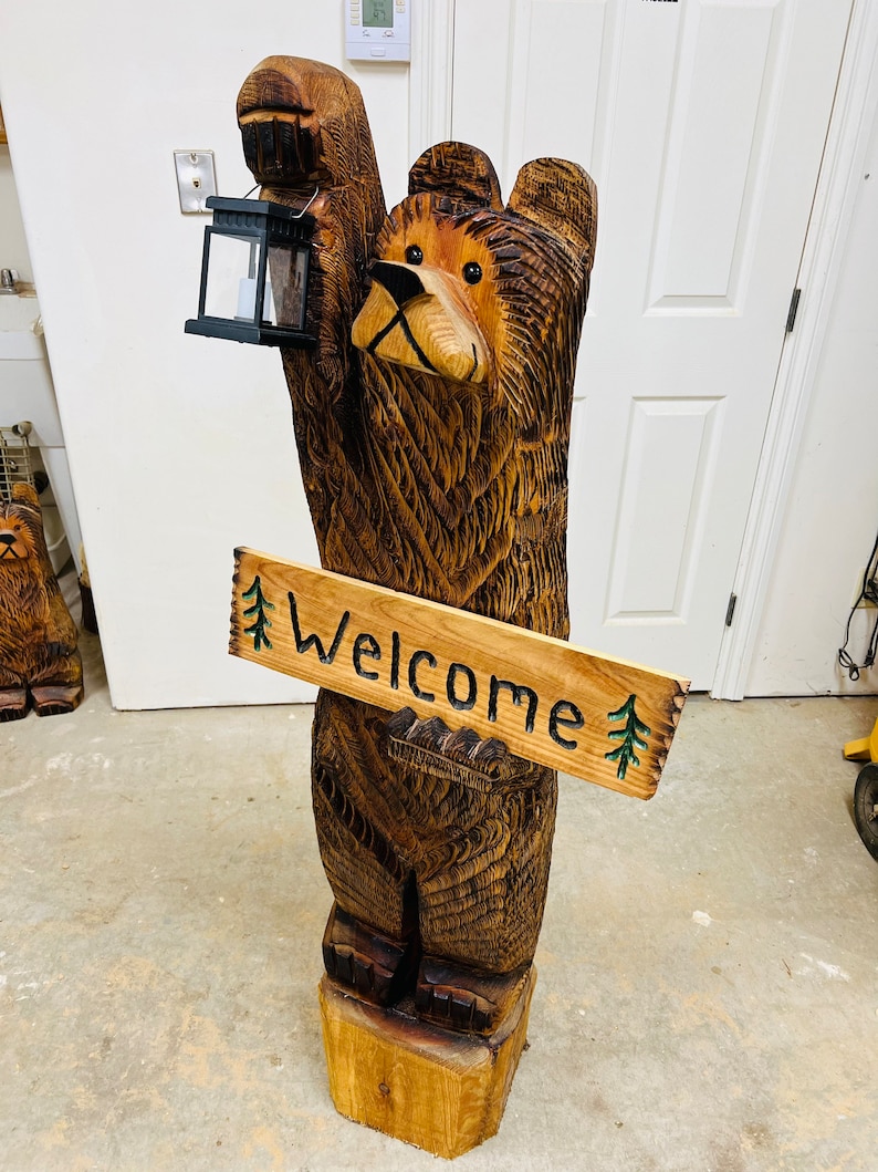 May include: A carved wooden bear statue holding a lantern and a sign that reads "Welcome." The bear is brown with detailed wood grain, standing on a wooden base. The sign has green painted trees.