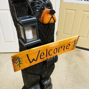 May include: A carved wooden black bear holding a sign that says "Welcome". The bear is holding a solar powered lantern in its other hand. The bear is standing on a wooden base.