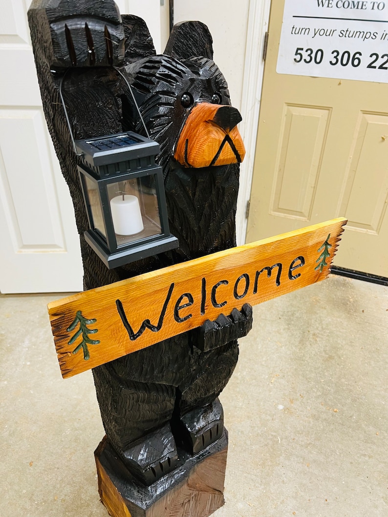 May include: A carved wooden black bear holding a sign that says "Welcome". The bear is holding a solar powered lantern in its other hand. The bear is standing on a wooden base.