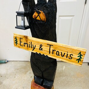 May include: A carved wooden black bear holding a lantern and a sign that reads "Emily & Travis" with green pine trees on either side of the text.