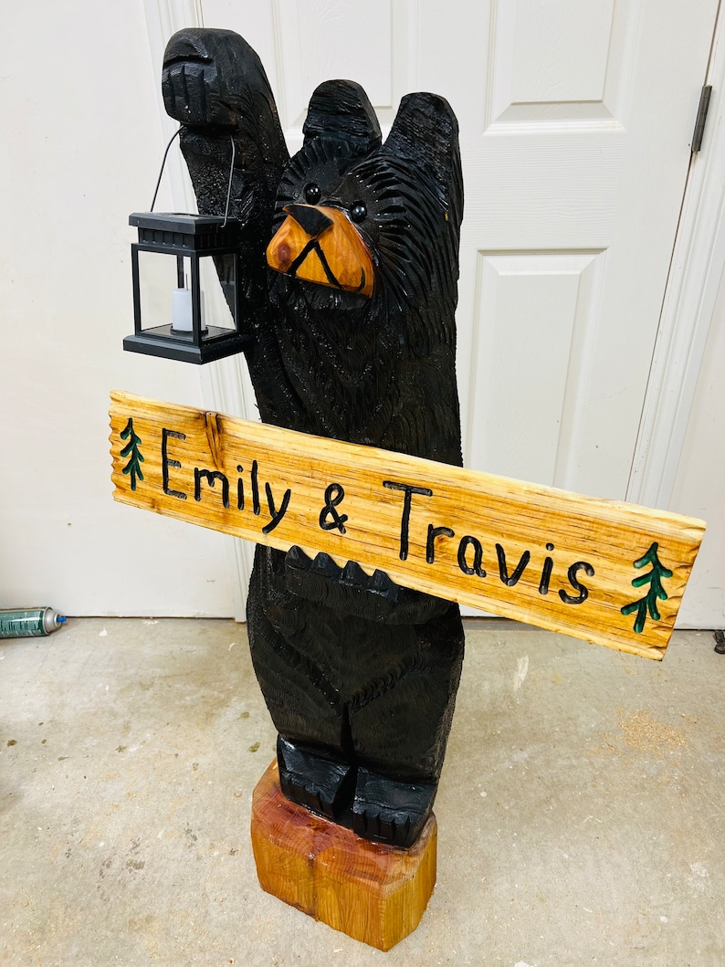 May include: A carved wooden black bear holding a lantern and a sign that reads "Emily & Travis" with green pine trees on either side of the text.