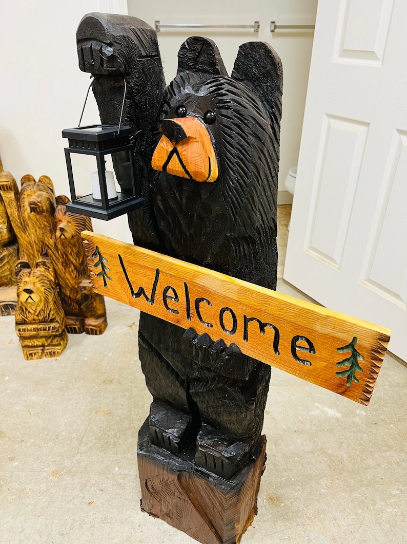 May include: A carved wooden black bear holding a lantern and a sign that says "Welcome". The bear is standing on a wooden base.