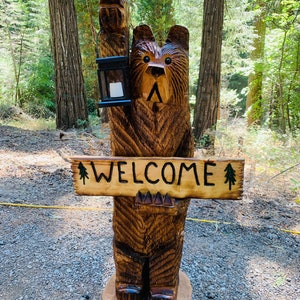 May include: A carved wooden bear sculpture holding a lantern and a sign that says "WELCOME". The bear is brown and has a friendly expression. The sculpture is standing on a wooden base in a forest setting.