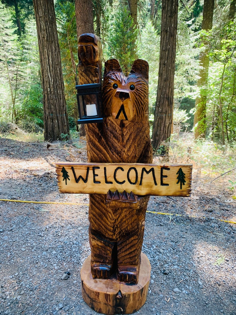 May include: A carved wooden bear sculpture holding a lantern and a sign that says "WELCOME". The bear is brown and has a friendly expression. The sculpture is standing on a wooden base in a forest setting.
