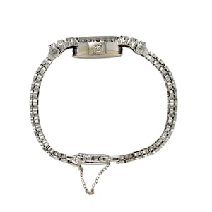 May include: A silver-toned wristwatch with a delicate, linked bracelet. The watch face is round, surrounded by small, sparkling stones. A small chain hangs from the clasp. The watch is designed for the wrist and is likely a vintage or antique piece.
