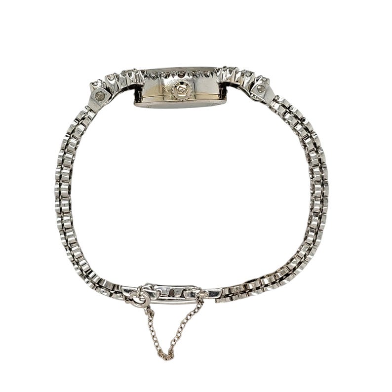 May include: A silver-toned wristwatch with a delicate, linked bracelet. The watch face is round, surrounded by small, sparkling stones. A small chain hangs from the clasp. The watch is designed for the wrist and is likely a vintage or antique piece.