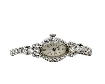 May include: An oval-faced Bulova watch with a silver dial and diamond-encrusted bezel and band. The watch features a small crown and is set against a white background. The watch hands are black.